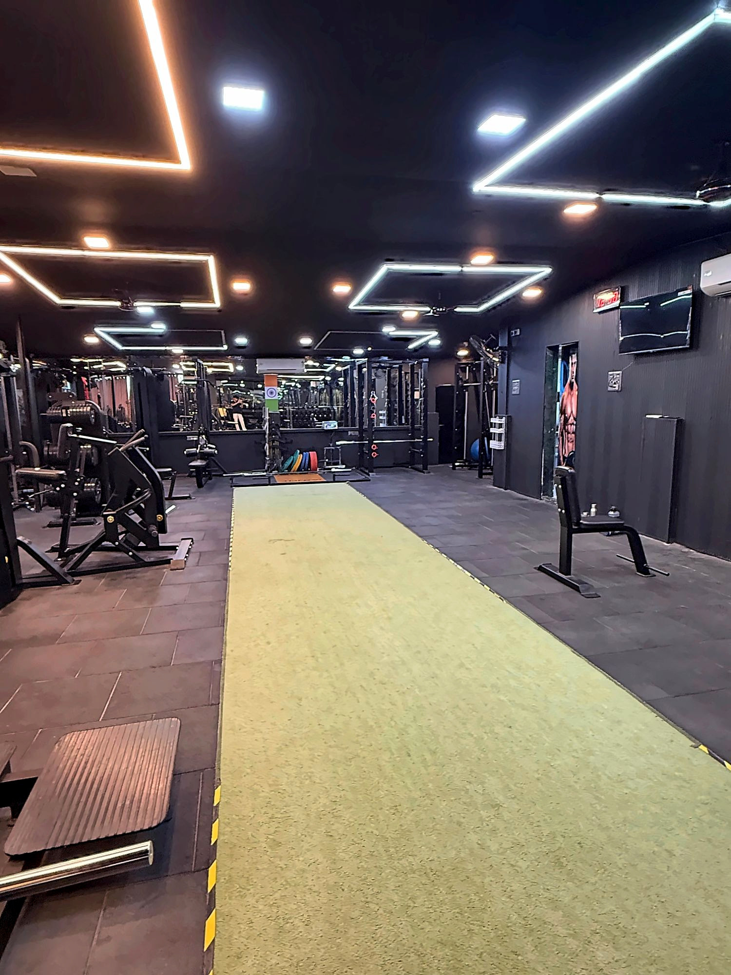 The EMPIRE GYM training floor: a turf lane between strength machines and racks under geometric LED ceiling lights